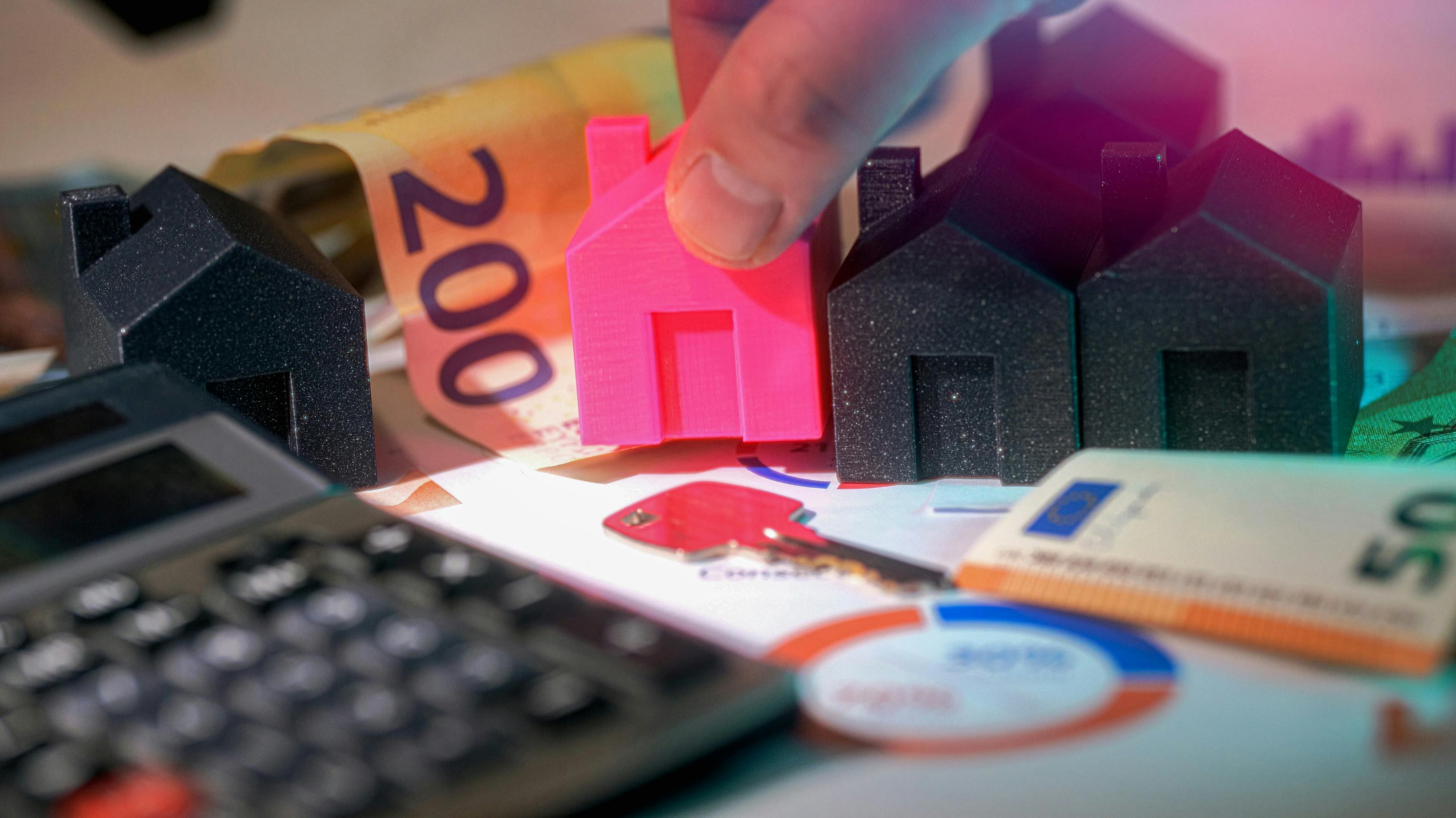 A close-up of a hand placing a small pink model house among three dark model houses on a desk strewn with euro banknotes, a calculator, a key and printed financial charts.